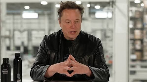 Elon Musk, de jaqueta de couro preta, com as mãos unidas diante do rosto, sentado a uma mesa com duas garrafas pretas, em um galpão