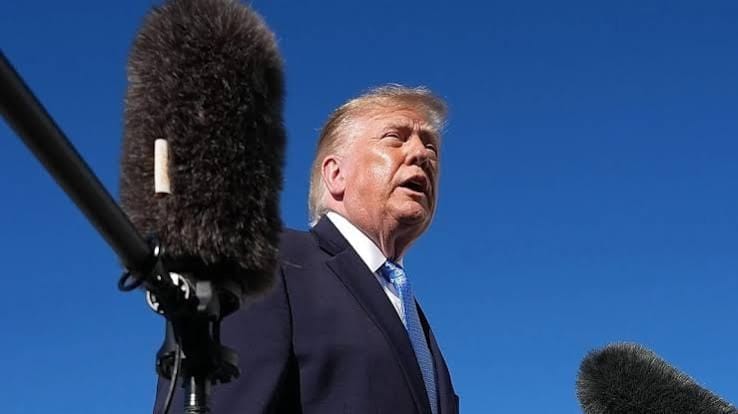 Donald Trump, de terno escuro e gravata azul, fala diante de microfones com protetor de espuma, com o céu azul ao fundo