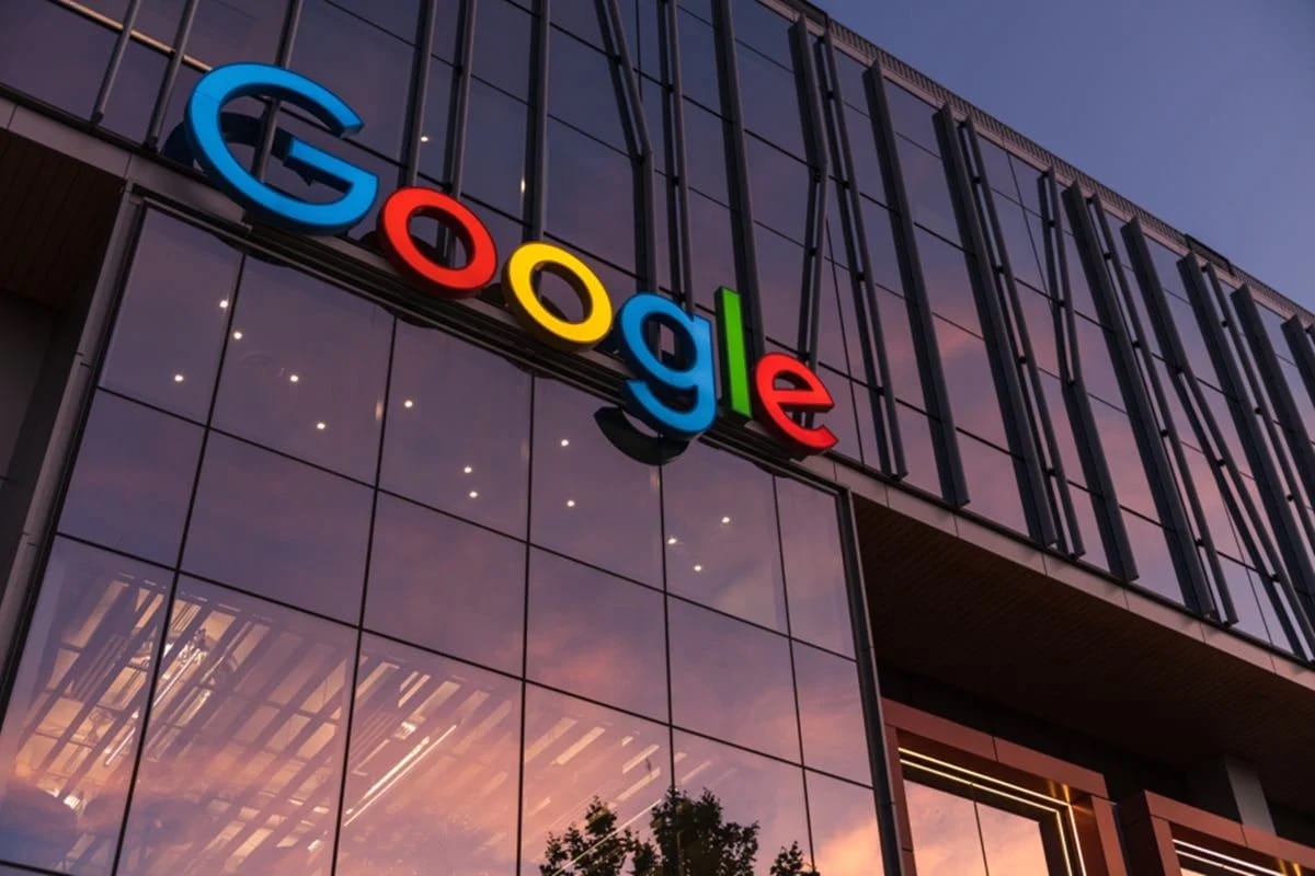 Fachada de vidro de um prédio corporativo com o logotipo colorido do Google ao entardecer, com o céu alaranjado refletido nas janelas