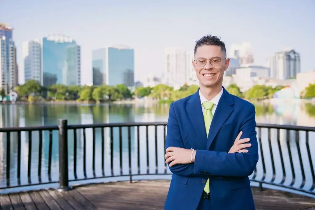 Homem de terno azul e gravata verde, de braços cruzados e sorrindo, posa em um deque de madeira com o lago e os prédios do centro de Orlando ao fundo
