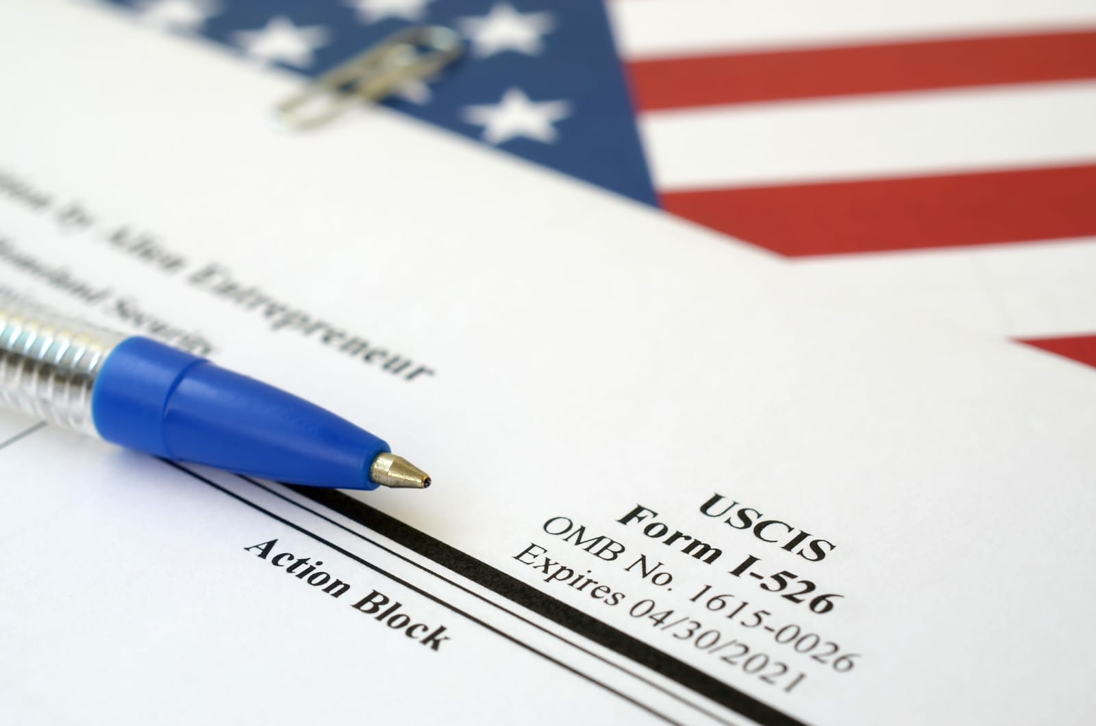 Formulário do USCIS sobre mesa, com caneta azul e bandeira dos Estados Unidos ao fundo