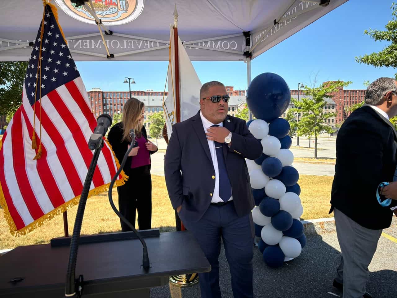 Homem de terno em evento público ao ar livre, com bandeira dos Estados Unidos e bandeira de Massachusetts ao fundo