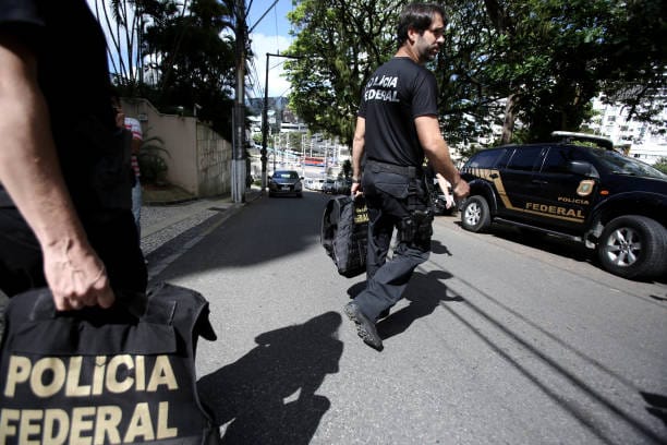 Agentes da Polícia Federal caminham pela rua durante operação, com colete escrito Polícia Federal