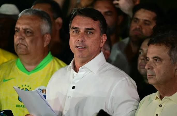 Flávio Bolsonaro, de camisa branca, fala segurando papéis, cercado por apoiadores, um deles de camisa amarela da seleção brasileira