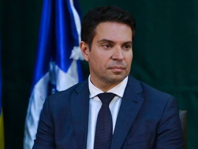 Alexandre Ramagem, de terno azul e gravata escura, sentado diante de uma bandeira azul e branca, com fundo verde