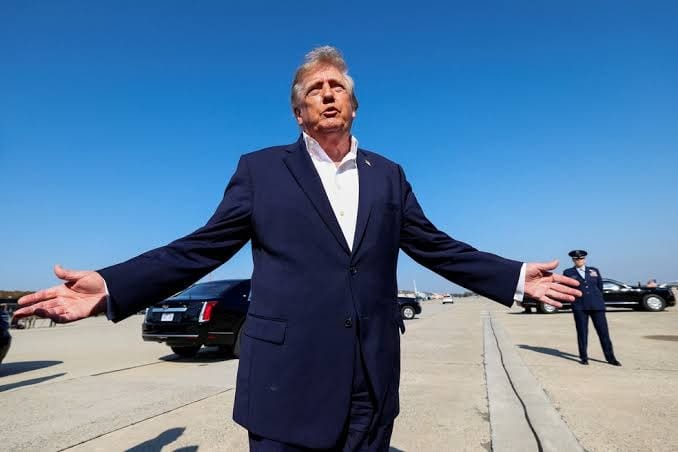 Donald Trump, de terno azul-marinho e camisa branca aberta, fala de braços abertos em uma pista, com carros pretos e um militar ao fundo