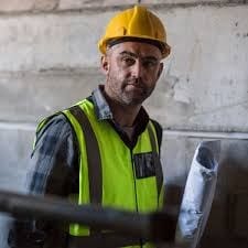 Trabalhador de capacete amarelo e colete refletivo verde segura rolos de papel em uma obra, diante de uma parede de concreto