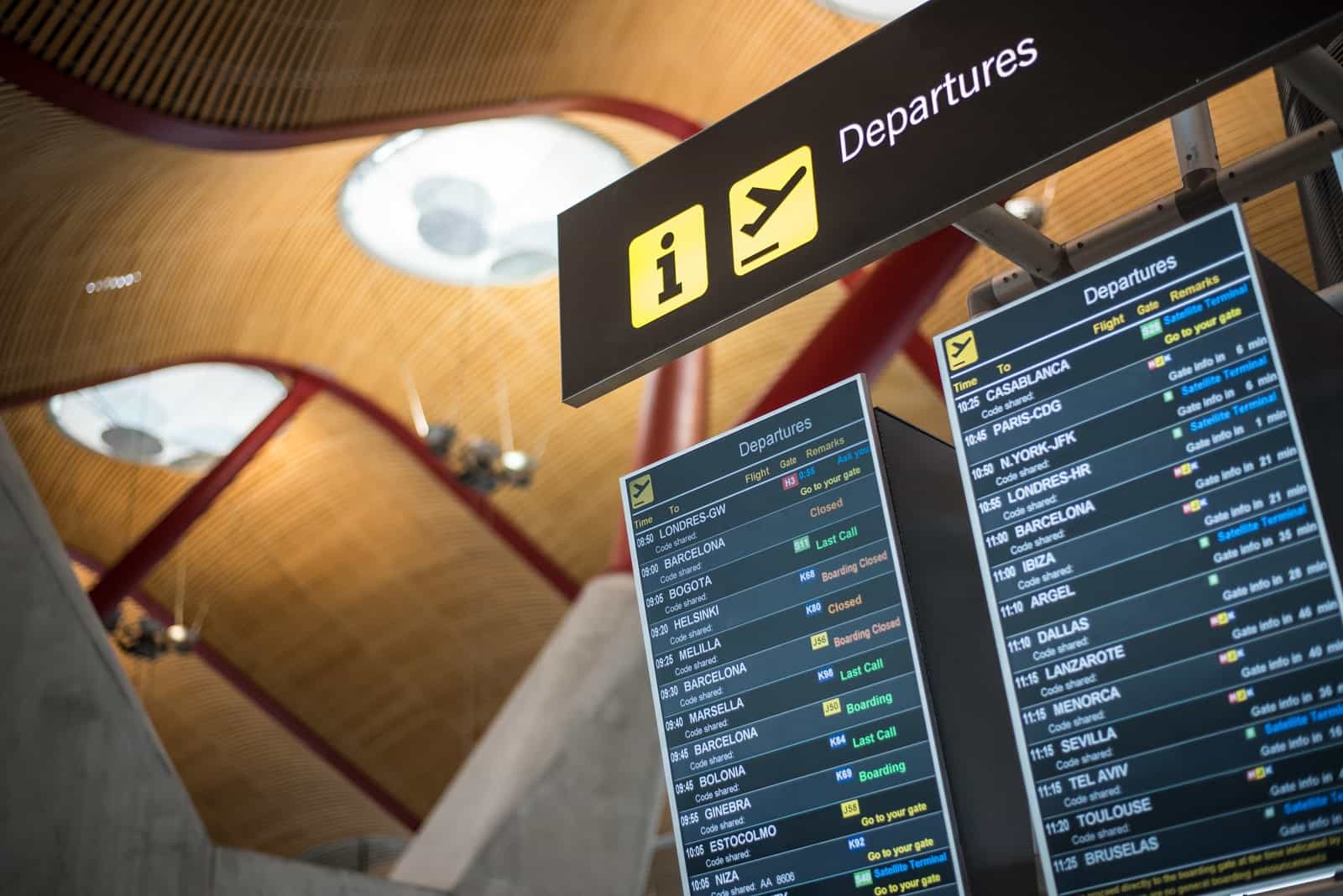 Placa Departures e dois painéis de embarque em um aeroporto, com voos para Londres, Paris, Nova York e outras cidades, sob um teto de madeira