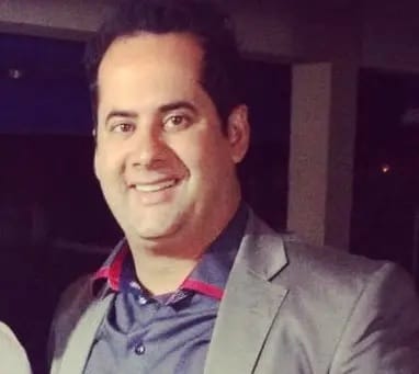 Retrato de um homem sorrindo, de cabelo escuro e blazer cinza sobre camisa social, em ambiente noturno