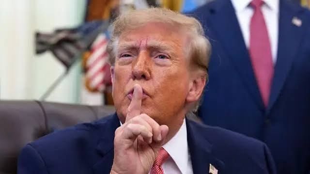 Donald Trump, de terno azul-marinho, faz o gesto de silêncio com o dedo sobre os lábios, em um gabinete com bandeiras ao fundo