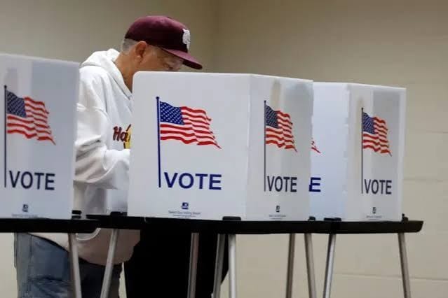 Eleitor de boné e moletom branco vota em uma cabine de papelão com a bandeira dos EUA e a palavra Vote, ao lado de outras cabines