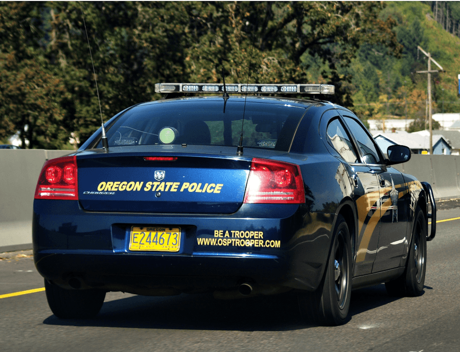 Traseira de uma viatura azul-marinho da Oregon State Police, com barra de luzes no teto e a inscrição OREGON STATE POLICE, rodando por uma rodovia