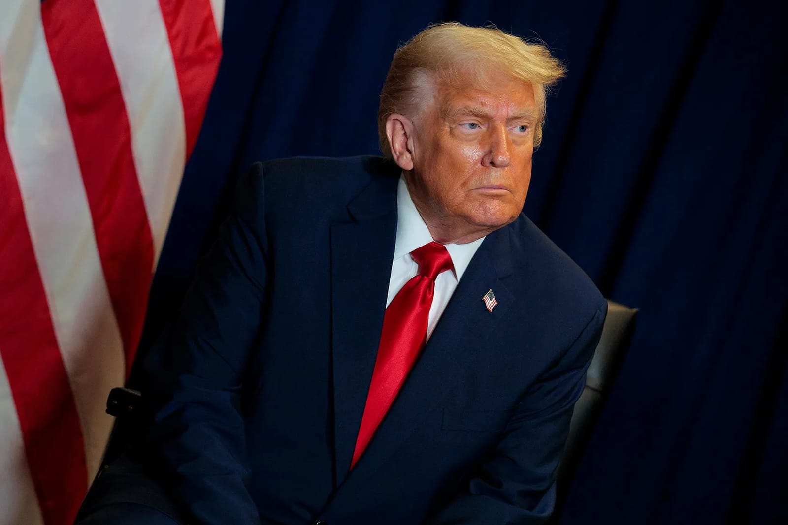 O presidente Donald Trump, de terno azul-marinho e gravata vermelha, sentado ao lado de uma bandeira dos Estados Unidos