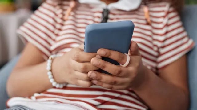 Close das mãos de uma criança, de camiseta listrada em vermelho e branco, segurando um celular com capa azul