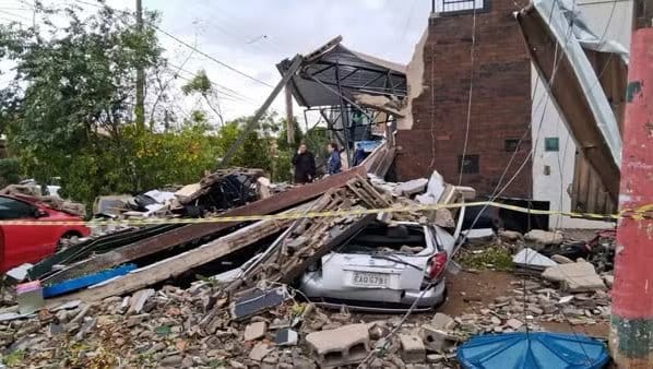 Destroços de uma estrutura desabada sobre carros, com escombros, fiação caída e fita de isolamento após um temporal; ao fundo, pessoas observam os estragos