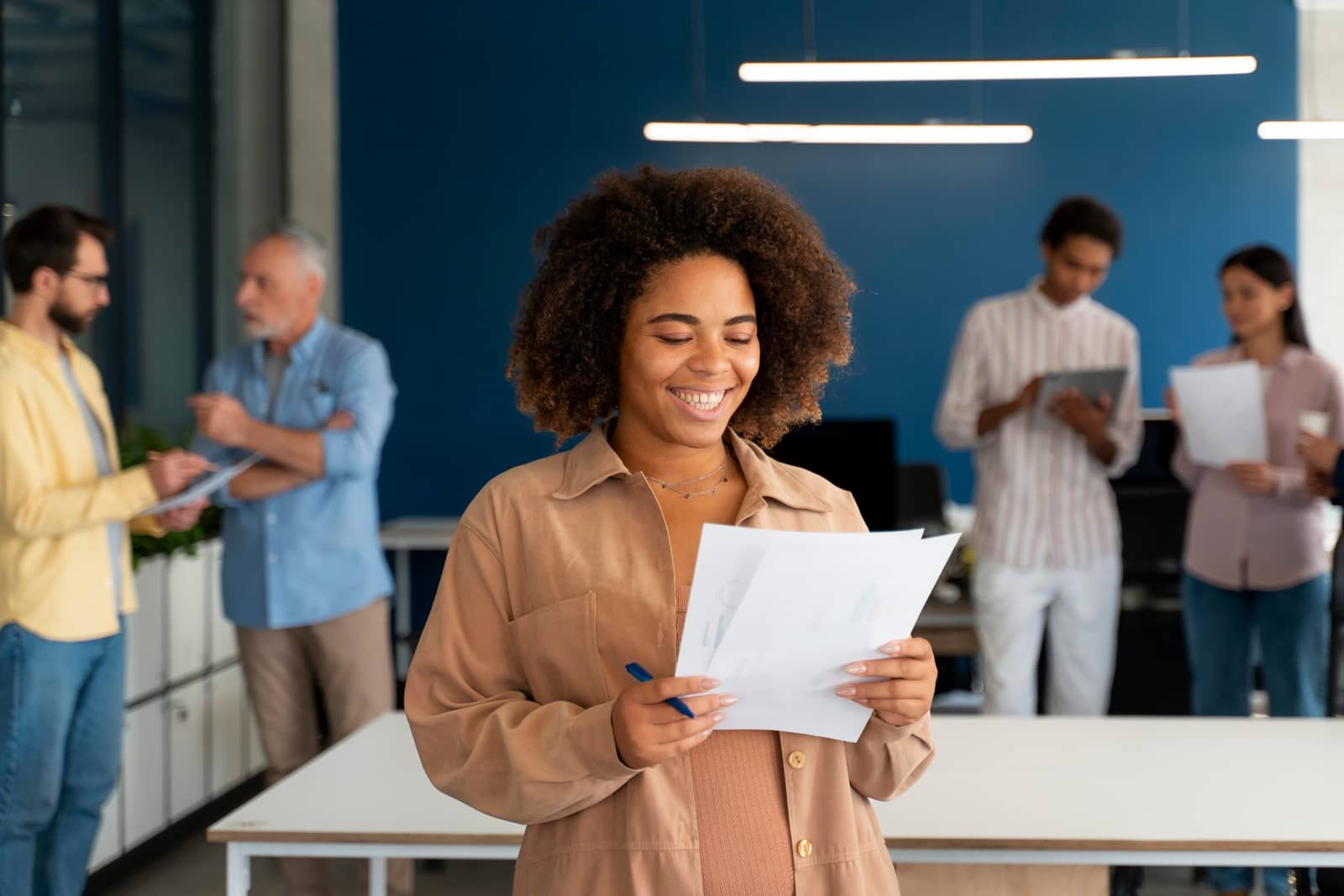 Mulher sorrindo com folhas de papel na mão em escritório, com colegas ao fundo