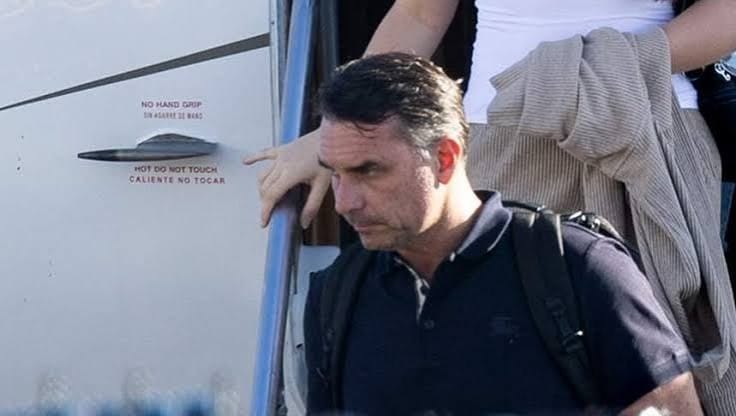 Homem de camisa polo escura e mochila desce a escada de um avião, apoiando a mão no corrimão, ao lado de uma porta com avisos em inglês e espanhol
