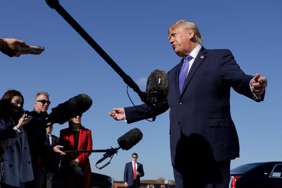 Donald Trump, de terno azul-marinho e gravata azul, fala de braços abertos diante de vários microfones e jornalistas, sob um céu azul