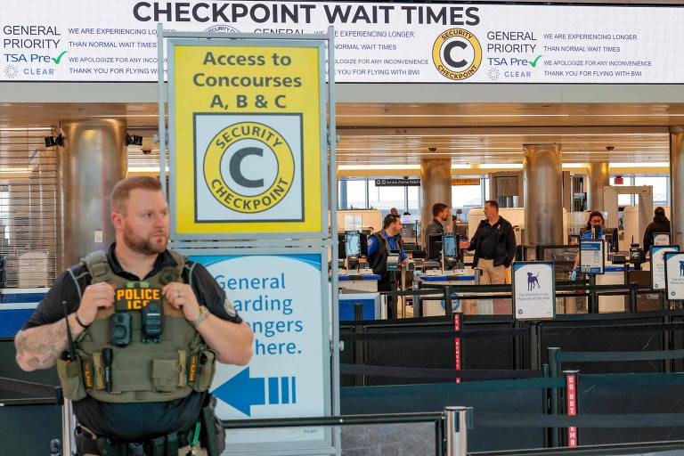 Agente com colete tático e a inscrição Police ICE em pé diante de um checkpoint de segurança de aeroporto americano, com painéis de tempos de espera ao fundo