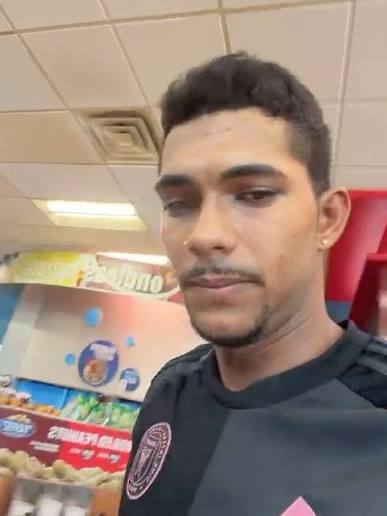 Homem jovem de cabelo escuro e camisa preta de time com detalhes rosa, em selfie dentro de um estabelecimento comercial
