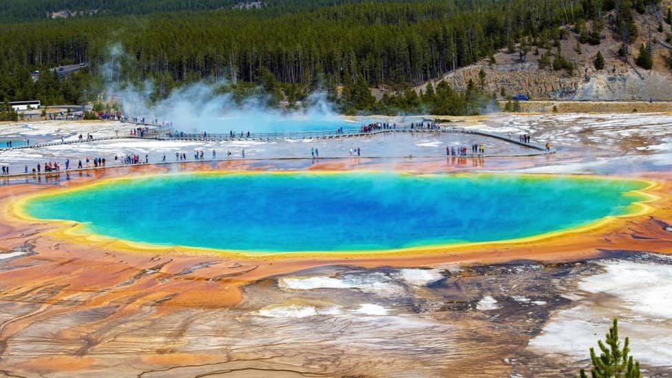 Vista elevada da Grand Prismatic Spring, em Yellowstone, com águas em tons de azul e bordas alaranjadas, vapor subindo e turistas caminhando pela passarela ao fundo
