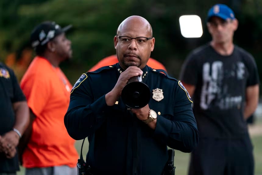 Shon Barnes, chefe de polícia de Seattle, de óculos e uniforme azul-marinho com distintivo, fala ao megafone em um espaço aberto, com pessoas ao fundo