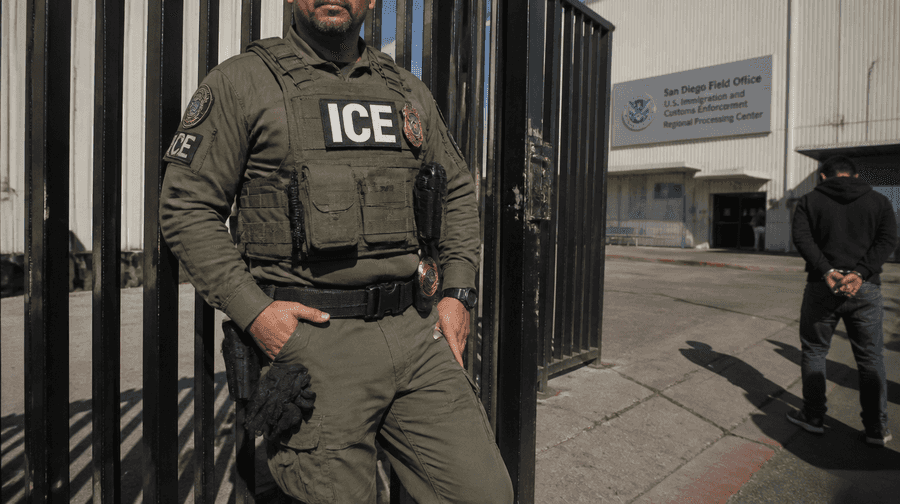 Agente do ICE em pé perto de um portão, com prédio da agência ao fundo e homem algemado à direita