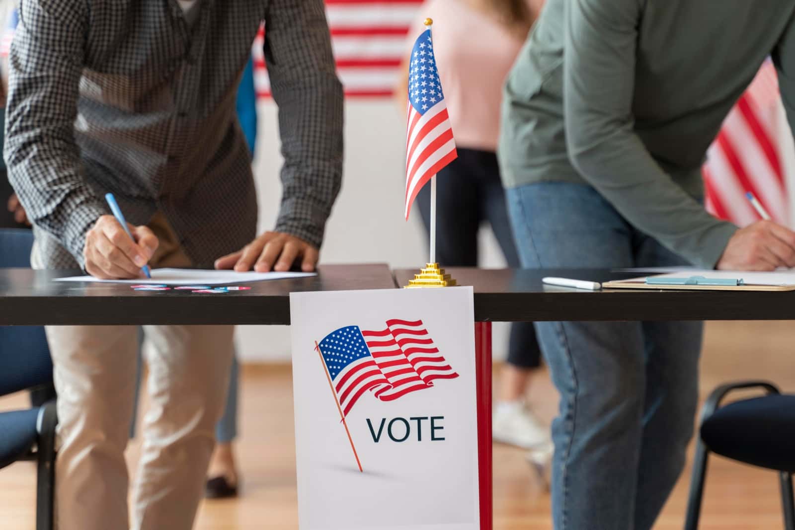 Eleitores preenchem formulários em uma mesa com uma pequena bandeira americana e um cartaz com a palavra Vote